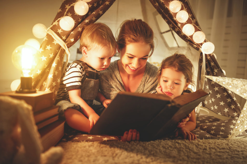happy family mother and children reading a book with a flashlight in a tent at home
