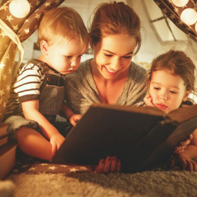 Eine Mutter liest ihren Kindern in einer gemütlichen, selbstgebauten Höhle mit Lichterketten aus einem Buch vor.