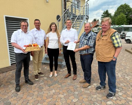Sechs Personen stehen in einem Halbkreis vor einem Gebäude und halten einen Vertrag in den Händen.
