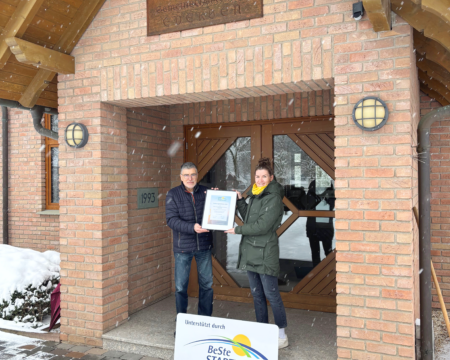 Ein Mann und eine Frau stehen im Eingang eines Gebäudes und halten eine Urkunde.