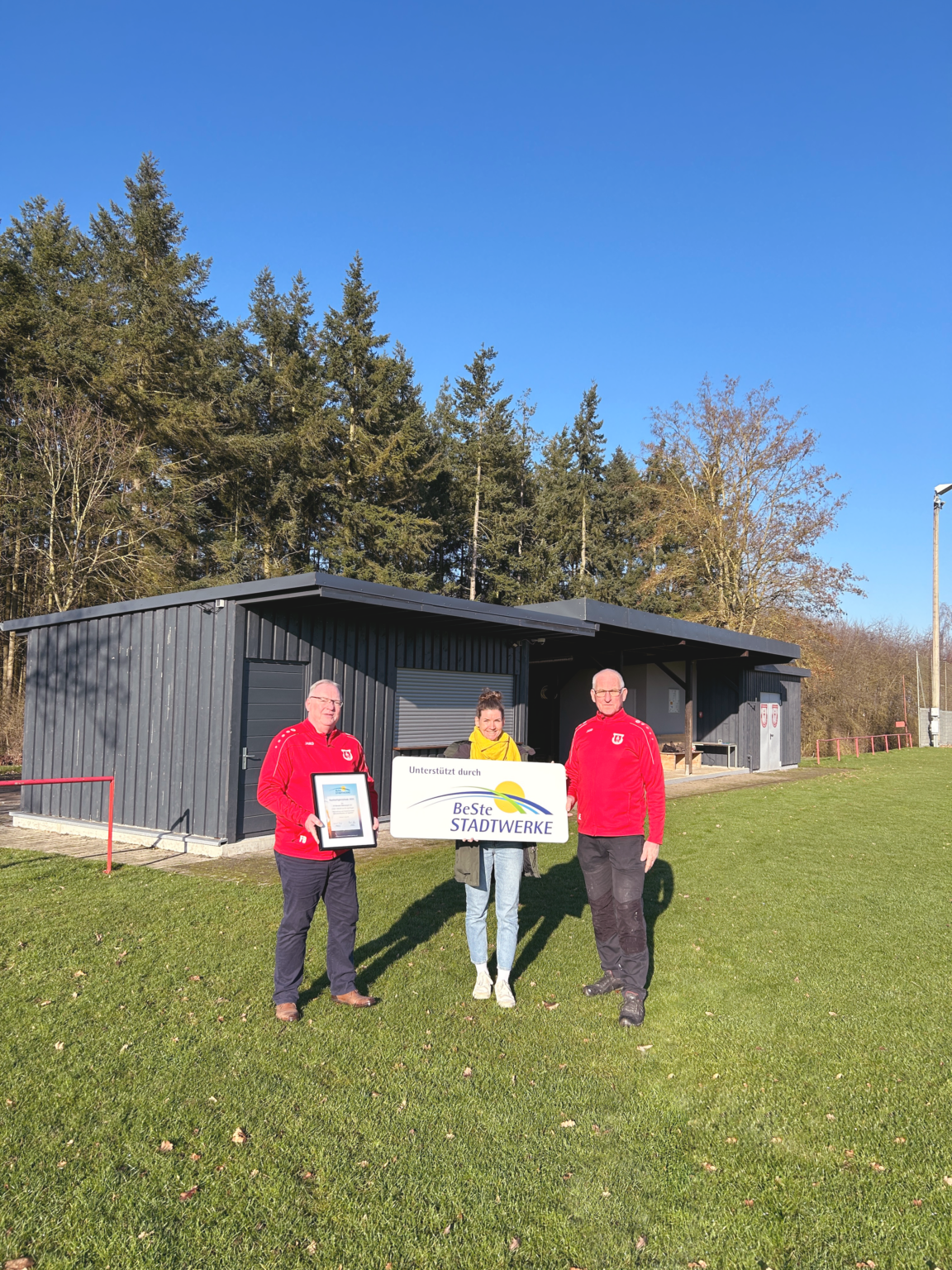Eine Frau und zwei Männer stehen mit Urkunde und Schild vor Vereinsheim.