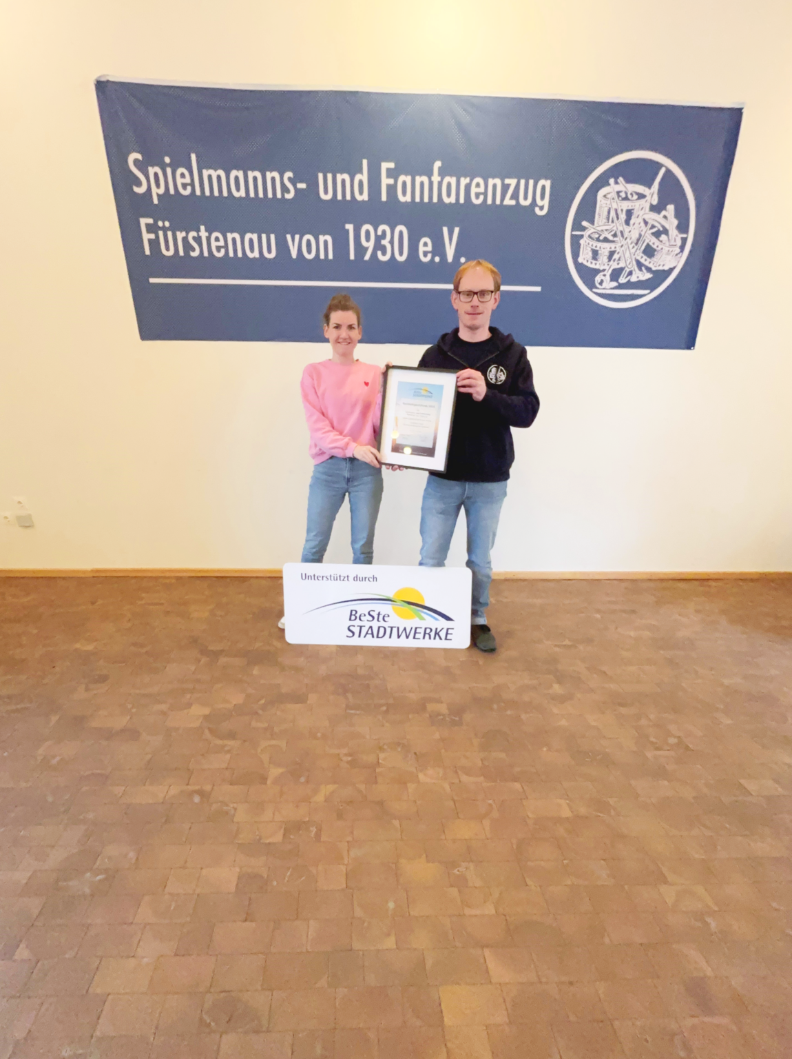 Eine Frau und ein Männer stehen mit Urkunde und Schild vor einem Banner des Vereins.