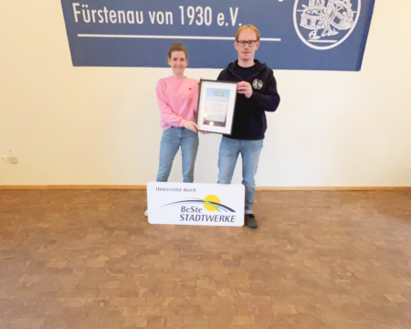 Eine Frau und ein Männer stehen mit Urkunde und Schild vor einem Banner des Vereins.