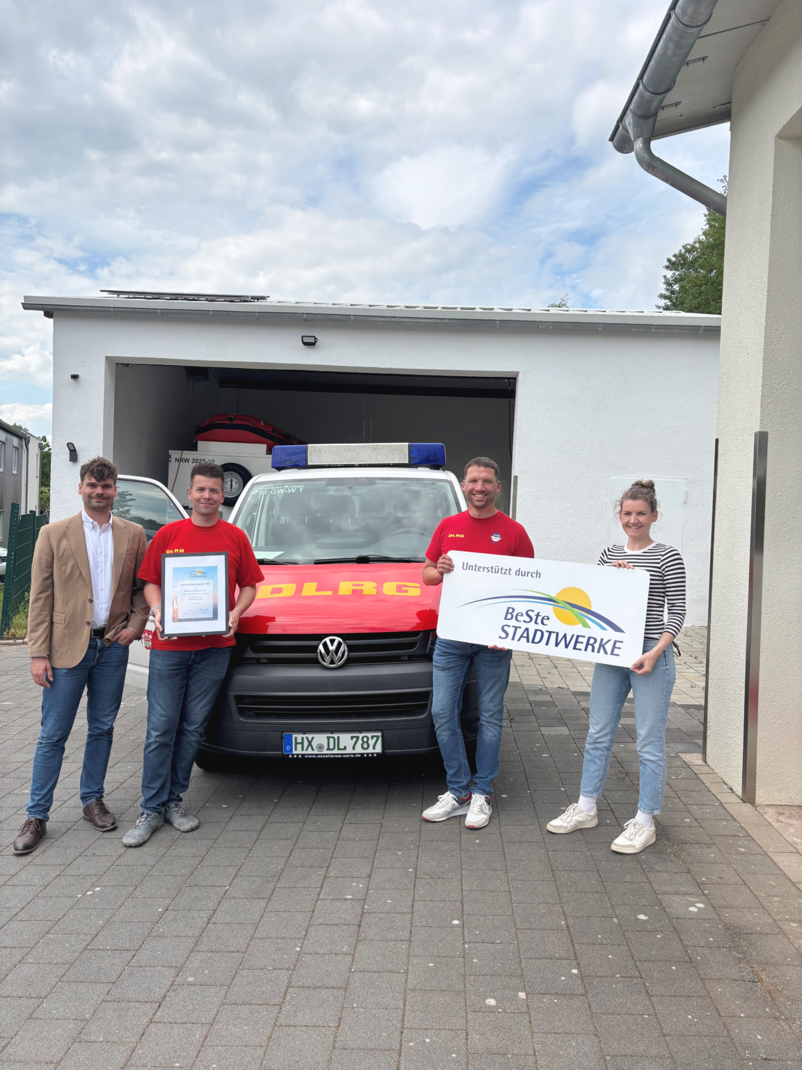 Vier Personen stehen vor einem DLRG-Einsatzfahrzeug in einer Garage und halten eine Urkunde sowie ein Schild „Unterstützt durch BeSte Stadtwerke“.