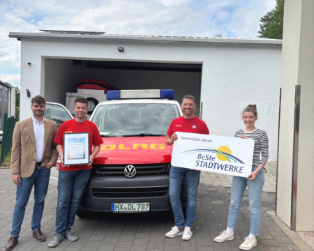 Vier Personen stehen vor einem DLRG-Einsatzfahrzeug in einer Garage und halten eine Urkunde sowie ein Schild „Unterstützt durch BeSte Stadtwerke“.