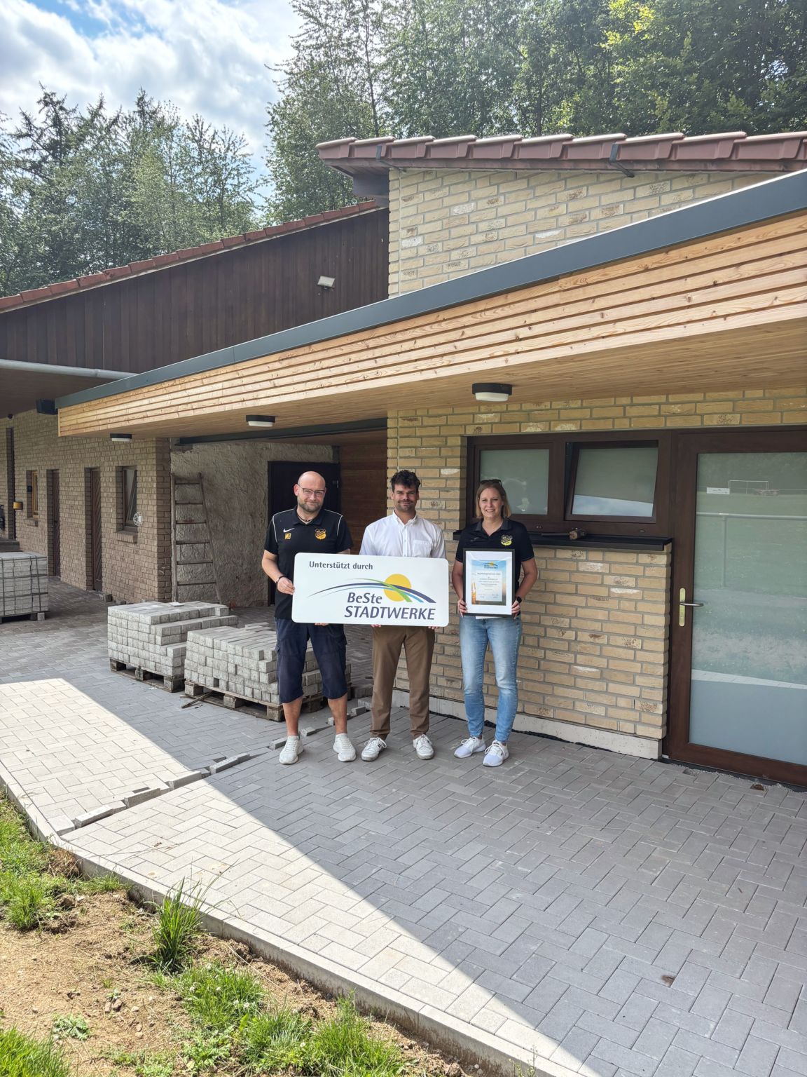 Drei Personen stehen mit Urkunde und Firmenschild vor Vereinsheim